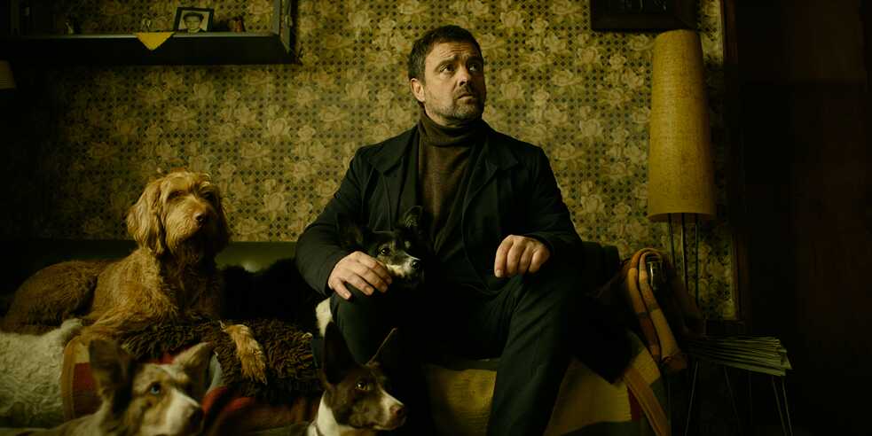 "Parfum - Ambra": Hauptkommissar Matthias Köhler (Jürgen Maurer) sitzt auf einer Couch. Neben, sowie vor ihm sitzen jeweils zwei Hunde, die in die gleiche Richtung wie er schauen.
