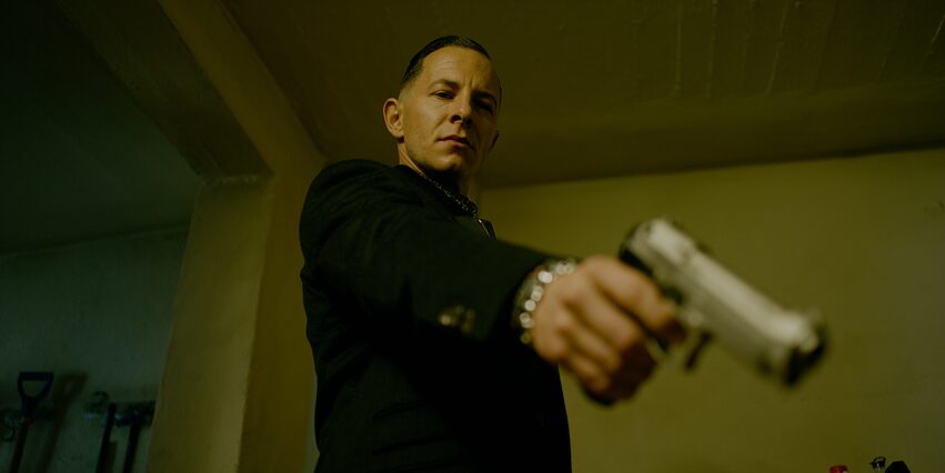 "Parfum - Fesselung": Unteransicht auf Thomas Butsche (Trystan Pütter). Er steht in einem Zimmer, mit ausgestrecktem Arm richtet er seine Waffe schräg nach unten in Richtung Kamera.