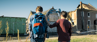 Two boys turning their back to the camera. One has a hand on the other one's shoulder while watching the demolition of a house.