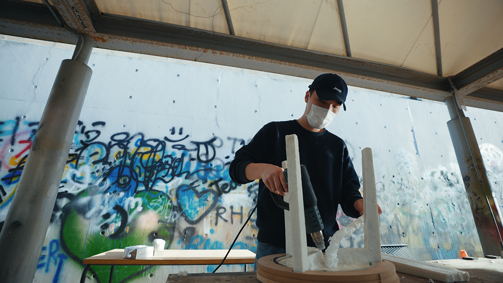 Haneul Kim is standing in front of a wall of graffiti and working on one of his chairs with a kind of heat gun