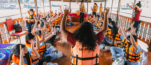 Young people kneel on the deck of a wooden boat and raise their arms. They wear life jackets.
