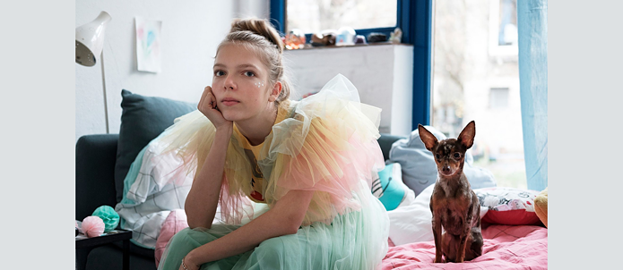 Une fille en costume coloré est assise sur le bord du lit, à côté d'elle se trouve un chien.