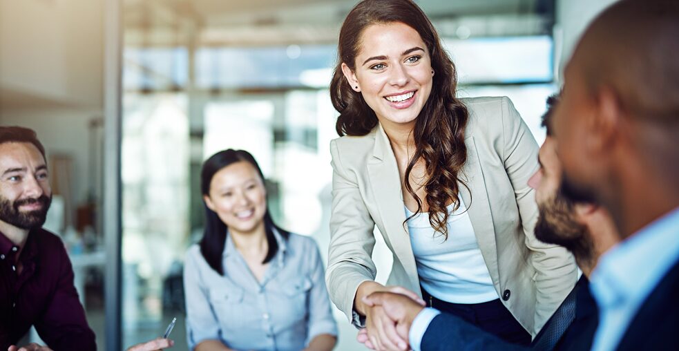 Eine junge Frau schüttelt lächelnd einem Mann die Hand, der mit anderen Menschen an einem Tisch sitzt.