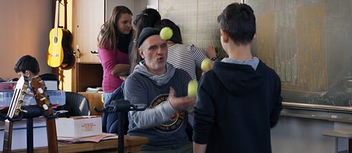 Scene from the film "Mr Bachmann and his class" – There are many guitars in the classroom. In the background three students are writing on the board. In the foreground a man is juggling three tennis balls. A student with his back towards the camera is observing him as he juggles.