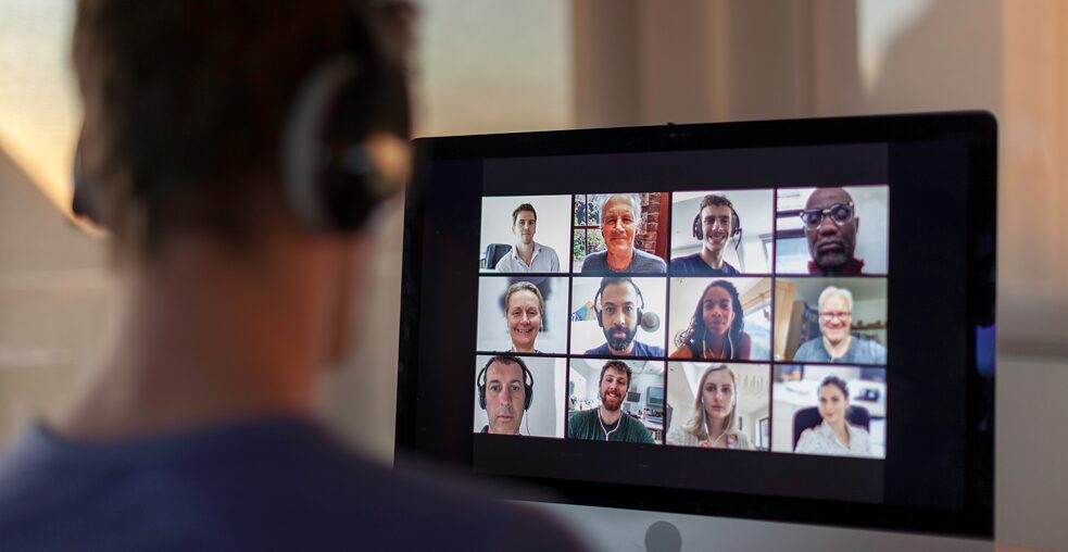 Man sieht einen Mann von hinten, der auf einen Bildschirm mit vielen Videokacheln sieht.