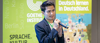 Male person with microphone, posters with inscription behind.