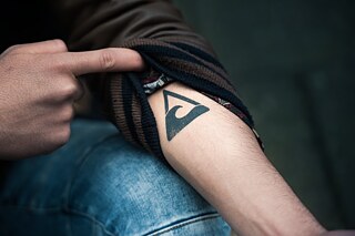 Standbild aus der Netflix Deutschland Serie „We are the wave/ Wir sind die Welle": Rahim zeigt sein Tattoo des Welle Logos