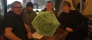 The four collaborators of the Walther von Goethe Foundation (from left to right: Wolfgang Müller, Ahmad Hamad, Richard Radzinski, An Paenhuysen) are sitting around a table, looking into the camera. In the centre there is a polygon shape, overgrown with moss. 