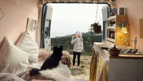 Doc Edge Festival 2022. Image from the film "Viral Dreams" showing the pink interior of a backpacker van including bed, sink, shelves, pictures and lights. There is also a small black dog on the bed in the van with the back doors open showing a  woman brushing her teeth and overlooking the forest landscape. 