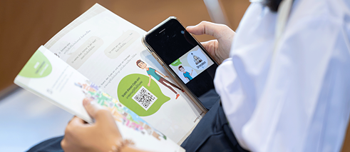 Eine Person hat das Buch „Kawin in Deutschland“ aufgeschlagen auf ihrem Schoß liegen. Sie hält ein Smartphone in der Hand, auf dem eine Animation abgespielt wird.