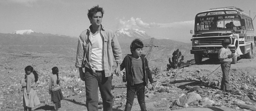 Vater nimmt Sohn an der Hand und geht mit ihm ein Weg in einer trockenen Berglanschaft entlang. Im Hintergrund sieht man einige Personen und einen alten Bus.