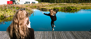 One young person filming another whose jumping in the air, next to a lake