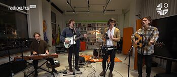A band consisting of four young men plays music in a room with a bookcase in the background.