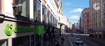 A street on which cars are driving. There are shops on both sides of the street, and on the left side in front you can see the Goethe-Institut.