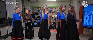 Five women with crowns sing into three standing microphones. Two of the women are holding violins. A bookshelf can be seen in the background.
