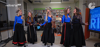Five women with crowns sing into three standing microphones. Two of the women are holding violins. A bookshelf can be seen in the background.