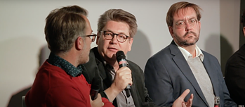 Three men with glasses, the middle one speaks into a microphone.