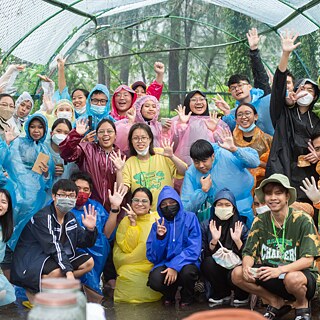A group of young people stand in nature and wave at the camera.