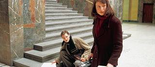 Sophie Scholl und ihr Bruder Hans Scholl beim Flyer verteilen in der Universität. Sophie Scholl im Vordergrund, ihr Bruder in der Hocke vor einem Treppenaufgang. Die beiden Schauen nervös in eine Richtung.