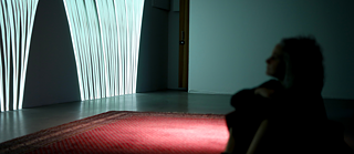 A woman in the blurry foreground, sitting in a dark exhibition space next to a red carpet and a large screen