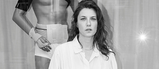 Black-and-white photograph, woman with long open hair and a white blouse is standing in front of a male mannequin.