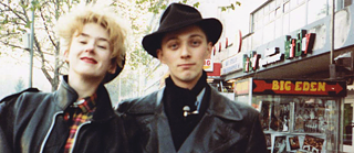 Man wearing a hat and woman on a street in Berlin, shops in the background