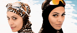 Close-up of the heads of two women in a swimming pool, both are wearing burkinis and swim goggles.