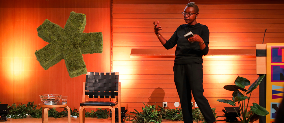 Neo Musangi on the Conference stage gesturing towards the audience (not in the picture). A moss asterisk floating in mid-air