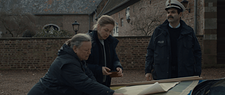 Polis üniforması giymiş bir adam ve iki kadın, polis arabasının üzerinde yerleştirilmiş büyük kağıtlara bakıyorlar. Arkalarında kırmızı bloklarla bir bina var.