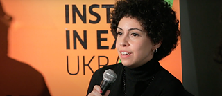 A woman with dark, curly hair speaks into a microphone.
