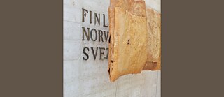 Foto einer hellen Betonmauer, die teilweise die Worte an der Wand in ihren eigenen Zeilen zeigt: "finl, norv, svez" - vermutlich in einer der Sprachen Finnisch, Norwegisch und Schwedisch. Oberhalb des Textes befindet sich eine Tasche oder Skulptur aus Birkenrinde.