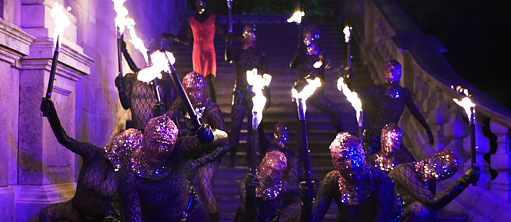 Personas disfrazadas bailan en una escalera con antorchas en las manos. La imagen irradia misterio y suspense.