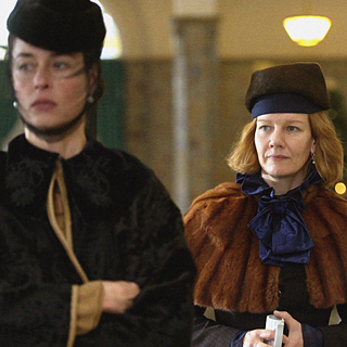 Zwei edel gekleidete Frauen laufen durch eine Eingangshalle.