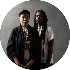 Black Quantum Futurism (Rasheedah Philips und Camae Ayewa) sind stehend vor einem grauen Hintergrund abgebildet. Sie schauen mit neutralen Gesichtsausdrücken in die Kamera.