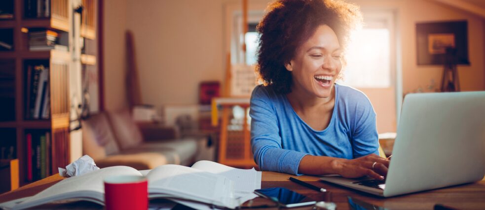 Eine junge Frau arbeitet an einem Laptop. Auf dem Tisch liegen Bücher, ein Handy und Notizen. Im Hintergrund sind Bückerregale, eine Couch und weitere Einrichtungsgegenstände unscharf zu sehen.