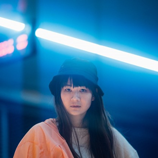 Artist Edy Fung is pictured looking directly into the camera. She is wearing a black hat and is standing in front of a dark, blurry background. A red neon sign and a blueish white neon lamp are visible in the background.