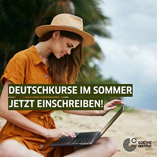 Eine junge Frau sitzt im Sand am Strand und schaut lächelnd auf ihren Laptop, sie trägt Sommerkleid und einen Hut
