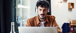 Un homme en situation d'apprentissage. Il est assis dans un café, porte des écouteurs et se concentre sur son ordinateur portable.