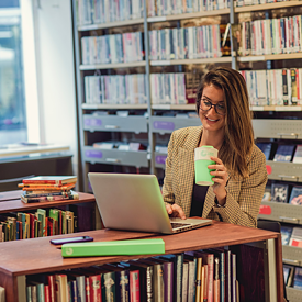 Eine junge Frau sitzt in der Bibliothek mit ihrem Laptop und einer Tasse Kaffe und lächelt.