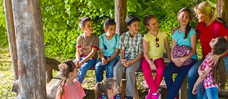 Una mujer rubia con coleta habla con un grupo de niños y niñas en un campamento