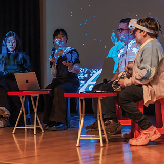 Four people sit on chairs on a stage and talk, a projection is visible in the background.