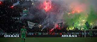 Ein Fußballspiel im Stadium. Auf der Tribüne wurden viele Feuerwerke gezündet. Es wehen Fahnen.