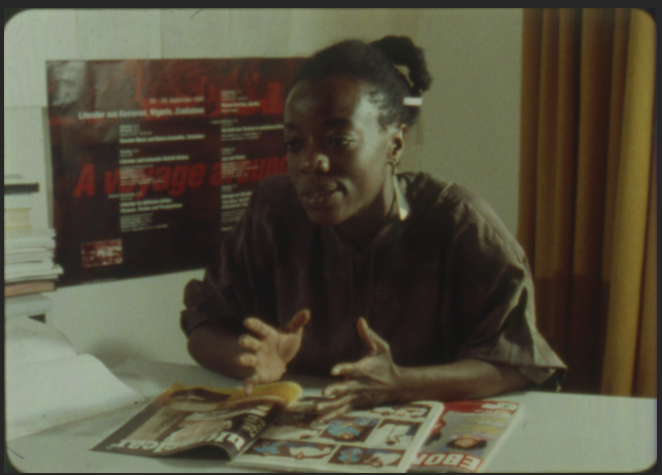 A balck woman sat at a table with old copies of Ebony opened before her. She is looking down, left of the camera and is using her hands to demonstrate