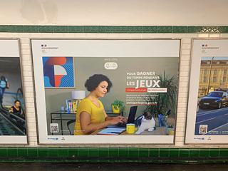 Ein Plakat in der Pariser Metro. Abgebildet ist eine Frau, die am Laptop arbeitet. Auf dem Schreibtisch sitzt eine Katze. Auf Französisch steht auf dem Plakat: "Um während der Spiele Zeit zu sparen, ist Telearbeit wichtig"
