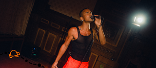 The Black artist LIE NING leans over a mirror laying on the ground and looks up at the camera. They are wearing a loose black latex top and golden earrings. In the right upper corner you can see a walking turtle that was drawn on the picture.