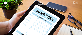 Eine Person hält ein Tablet, auf dem ein Bewerbungsformular angezeigt wird. Auf dem Schreibtisch liegen ein Notizbuch, ein Stift, eine Brille und ein Smartphone, was den digitalen Bewerbungsprozess symbolisiert.