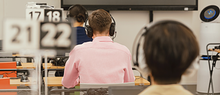 Mehrere Personen, die Kopfhörer tragen und an nummerierten Tischen in einem Klassenzimmer oder Testzentrum sitzen.