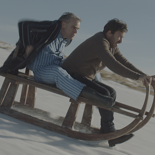 Billedet viser en ældre og en yngre mand på en slæde, der kører ned ad et bjerg i høj fart.