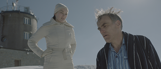 Das Bild bildet eine Szene aus dem Film ab. Es sind zwei Personen zu erkennen. Eine jüngere Frau mit weißen Mantel, Handschuhen und Mütze schaut von rechts herab auf einen Mann. Der Mann hat einen Schlafanzug unter einem Bademantel an und schaut entkräftet zu Boden.