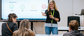 Eine Lehrerin mit blonden Haaren steht vorne im Unterrichtsraum des Goethe-Instituts Irland; sie lächelt die vier vor ihr sitzenden Schüler ermutigend an und streckt die Hand aus, um sie zum Antworten einzuladen. Auf einem Whiteboard im Hintergrund sind nützliche deutsche Wörter zu sehen.